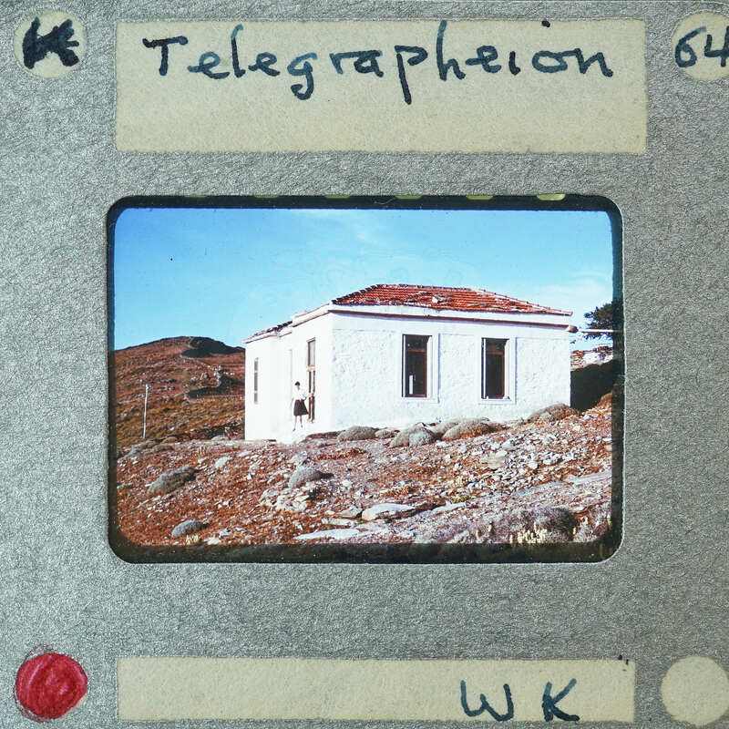 A vintage photograph of a small stone house with a red roof, surrounded by rocky terrain and sparse vegetation.