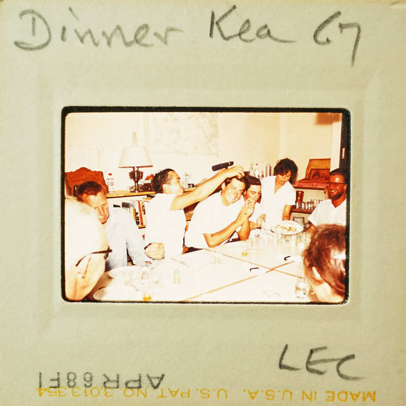 A vintage photograph of a group of people sitting around a table, possibly at a dinner party or gathering.