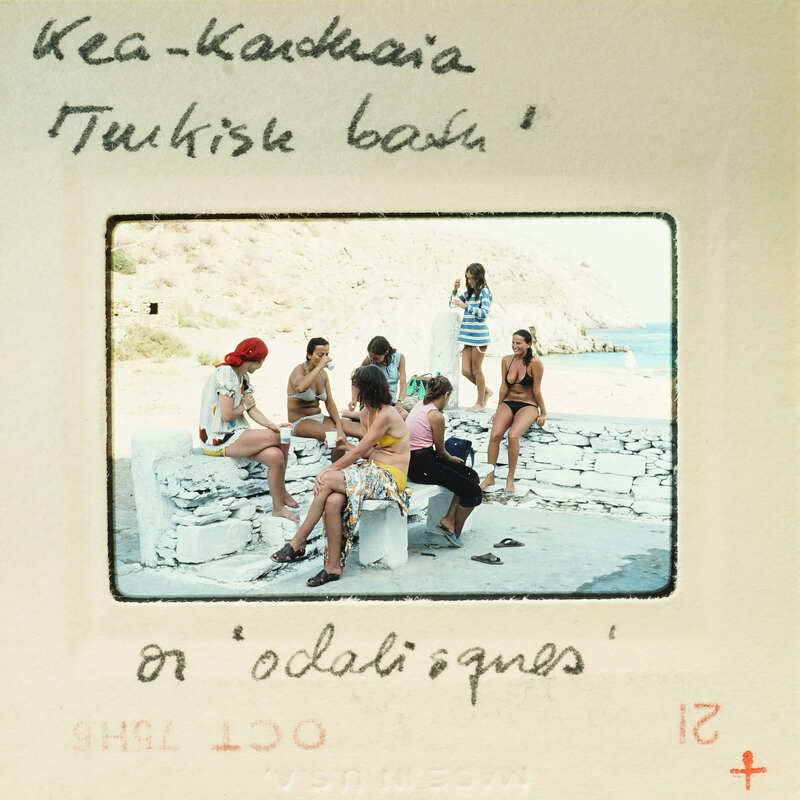 A vintage photograph of a group of people sitting on rocks by the water, with handwritten text and marks indicating it's from a film roll.