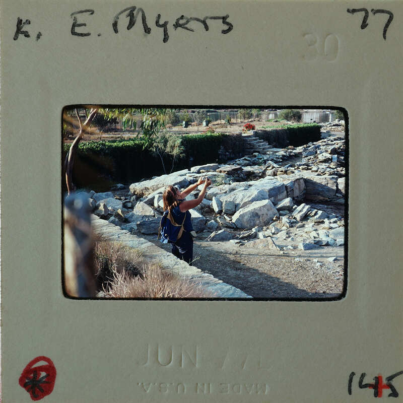 A person taking a photograph of rocks and trees, with the date "June 1977" visible on the slide frame.