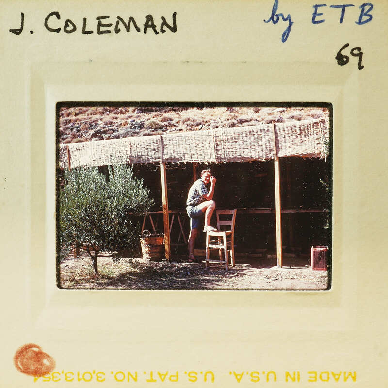 A vintage photograph of a man sitting on a stool under an outdoor shelter, with a plant and a chair nearby.
