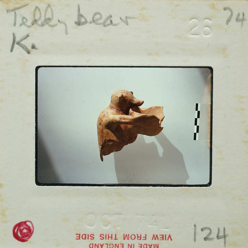 A photograph of a clay figurine with a person's hand visible, placed on an object identification card.