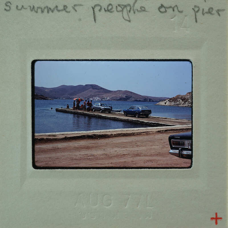 A vintage photograph of a group of people standing on a pier with a body of water and mountains in the background.