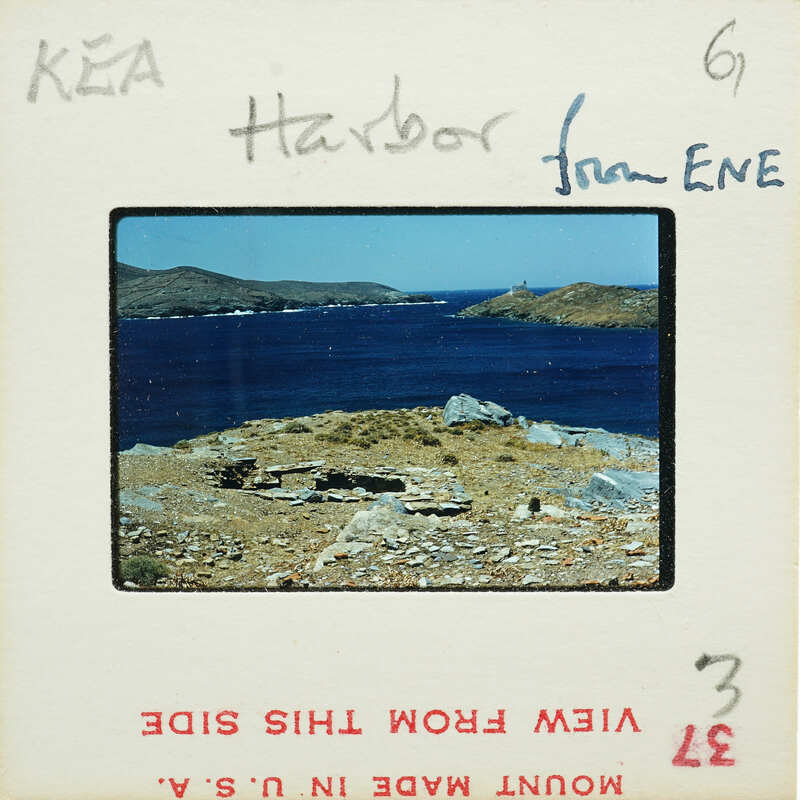 The image is a photograph of a scenic harbor with blue water, rocky shoreline, and distant islands under a clear sky.