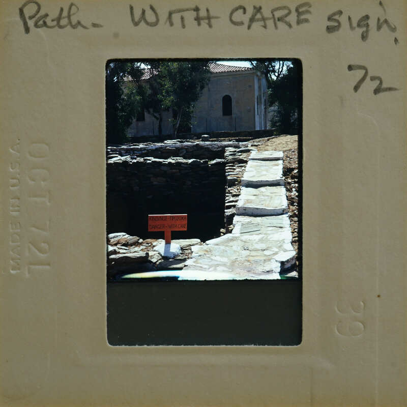 An old photo of a stone well with a sign on top, surrounded by ruins and greenery.