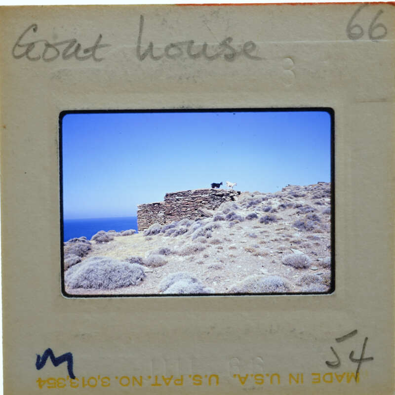 A photograph of a stone structure on a hillside with vegetation and the ocean in the background.