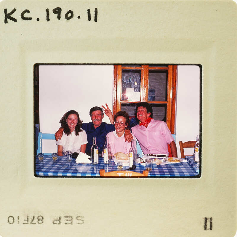 A vintage photo of a family posing together at a table with food and drinks, set against a window backdrop. Catalog number KC.190.11
