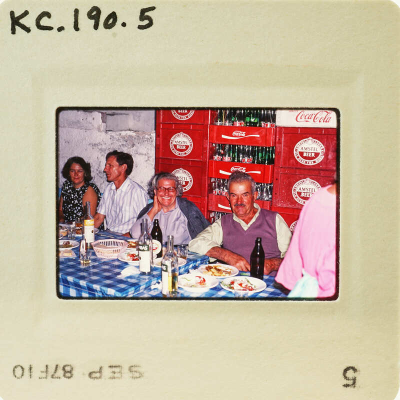 A vintage photo of people enjoying a meal together at a restaurant, with Coca-Cola crates visible behind them. Catalog number KC.190.5.