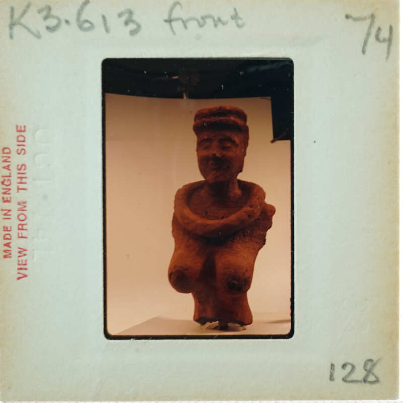 A photograph of a clay figurine with a smiling face and arms crossed, displayed on a white surface.