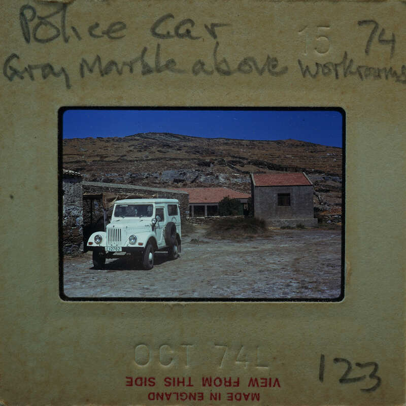 Old police car parked outside a village with mountains in the background.