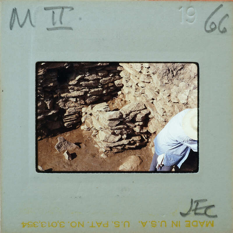 A person wearing a hat and blue shirt standing next to a stone wall with a brick pattern.
