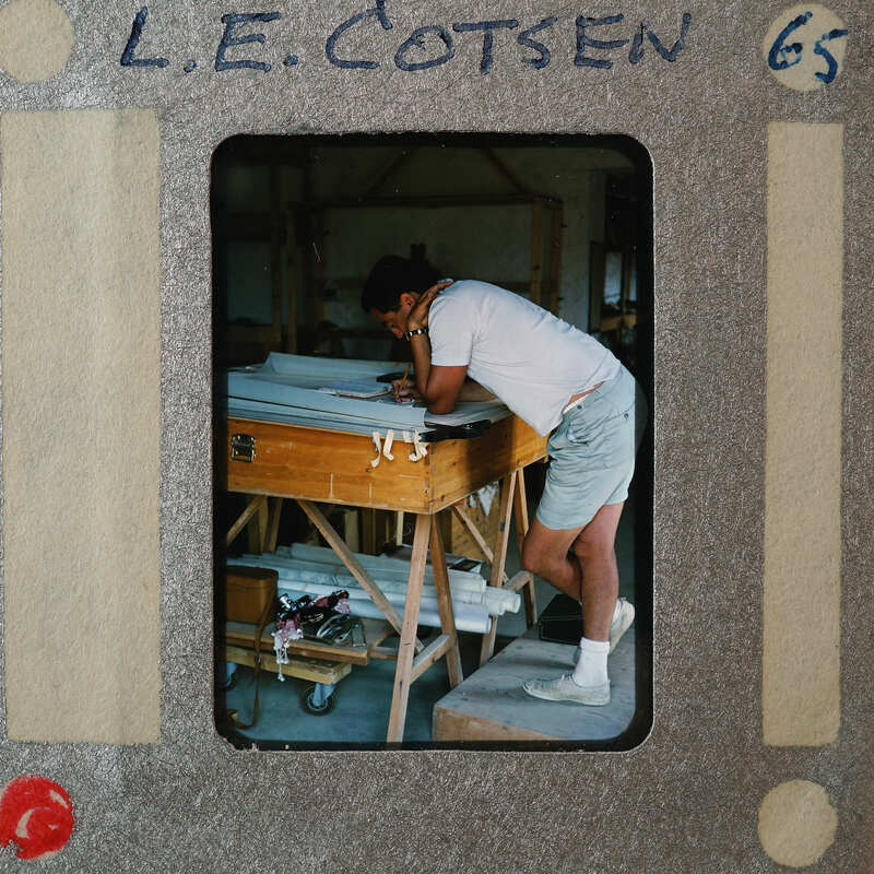 Man working at a table with various tools and materials around him, possibly in a workshop setting.