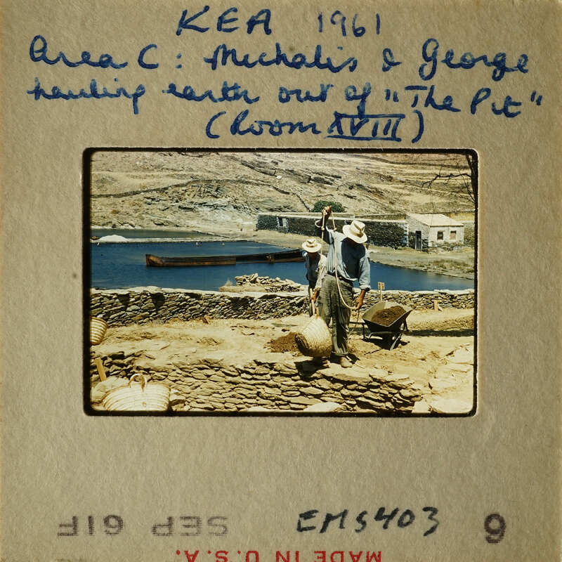Man shoveling dirt near a body of water with a boat and mountains in the background.