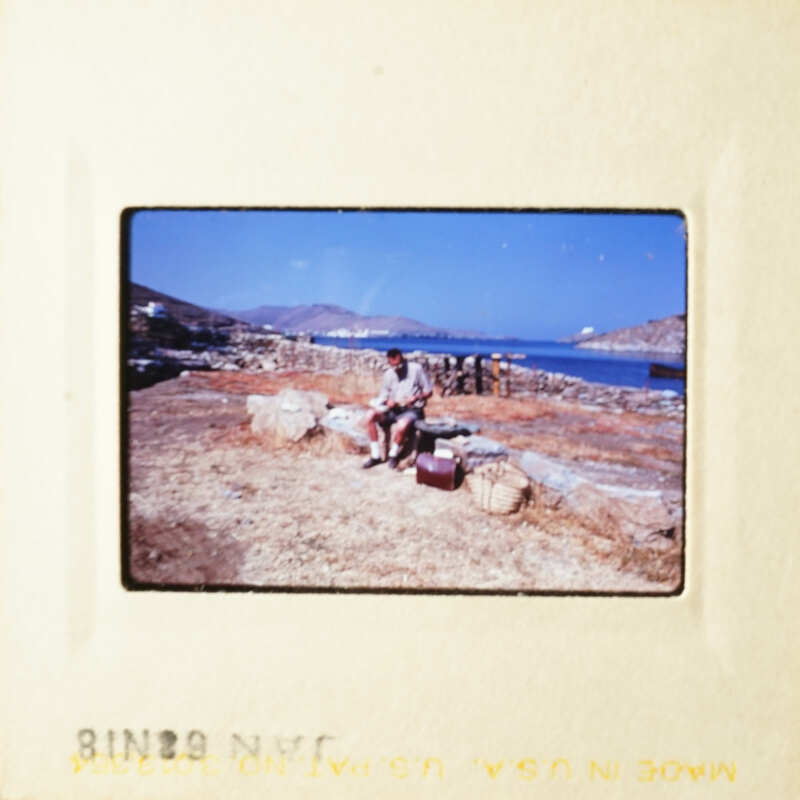 A man sitting on rocks next to the ocean, with mountains in the background.