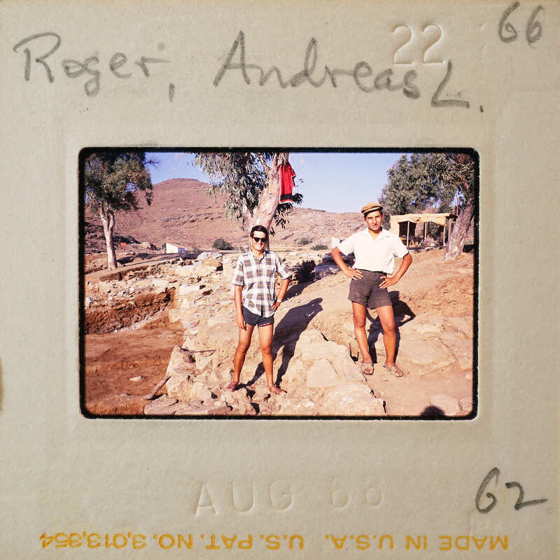 Two men standing on a rocky surface with mountains and trees in the background.