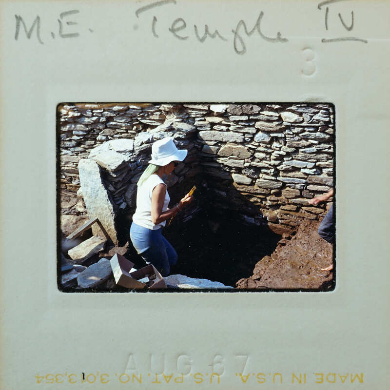 A person wearing a hat and a white shirt, standing next to a stone structure with a hole in the ground.