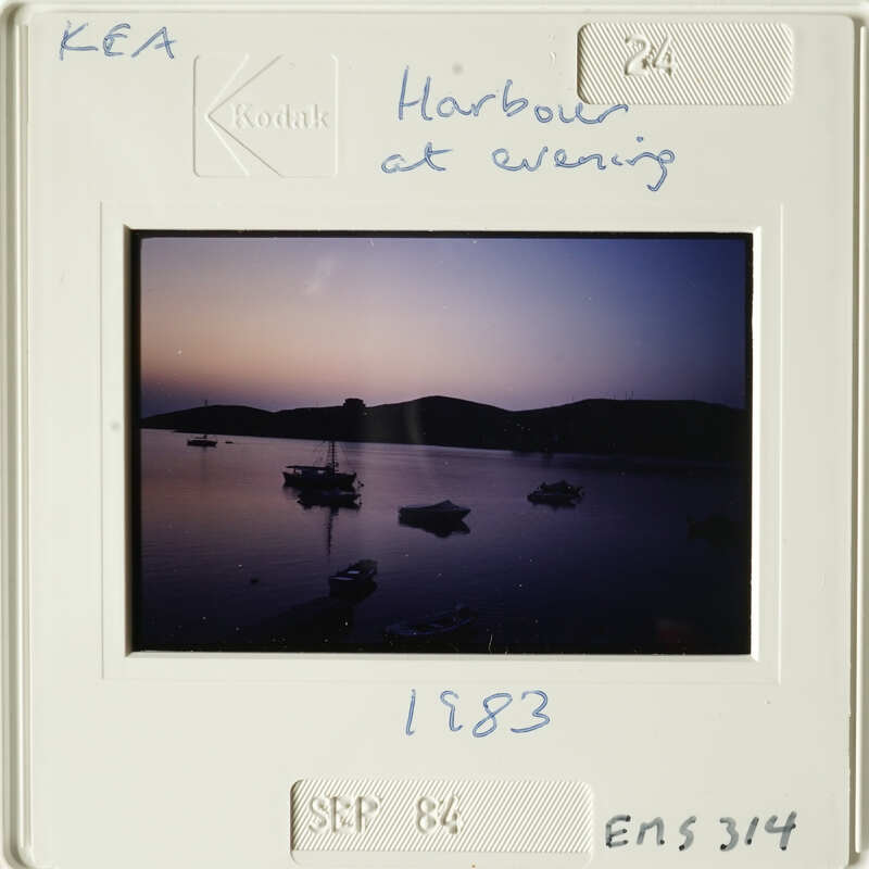 Harbor at evening with boats on water and mountains in background, captured in sepia-toned photograph.
