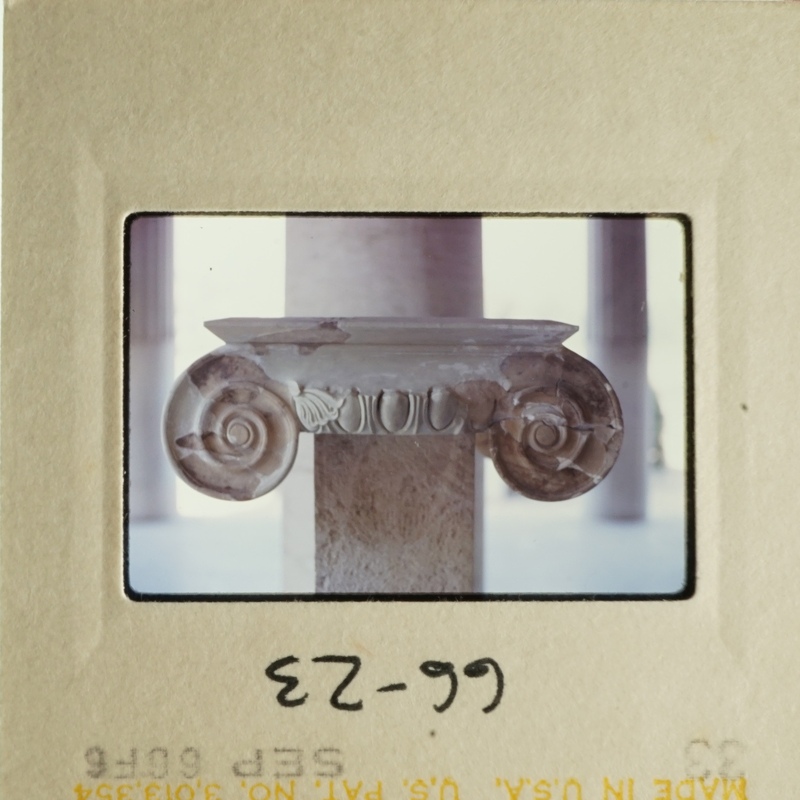 The photograph shows an ancient stone ionic column with decorative capitals and scrollwork, set against a plain background.