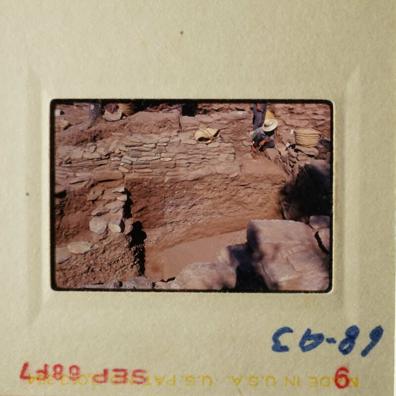 An archaeological site with stone structures, people working on excavation, and a dirt pathway.