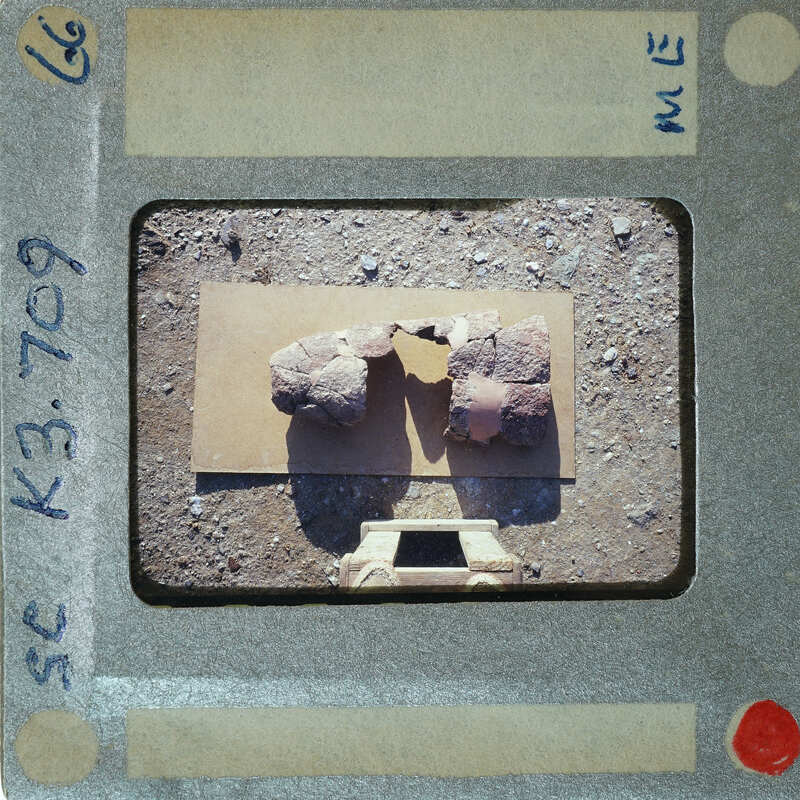 A photograph of an archaeological site with stone ruins and a measuring tool, taken from behind the camera.