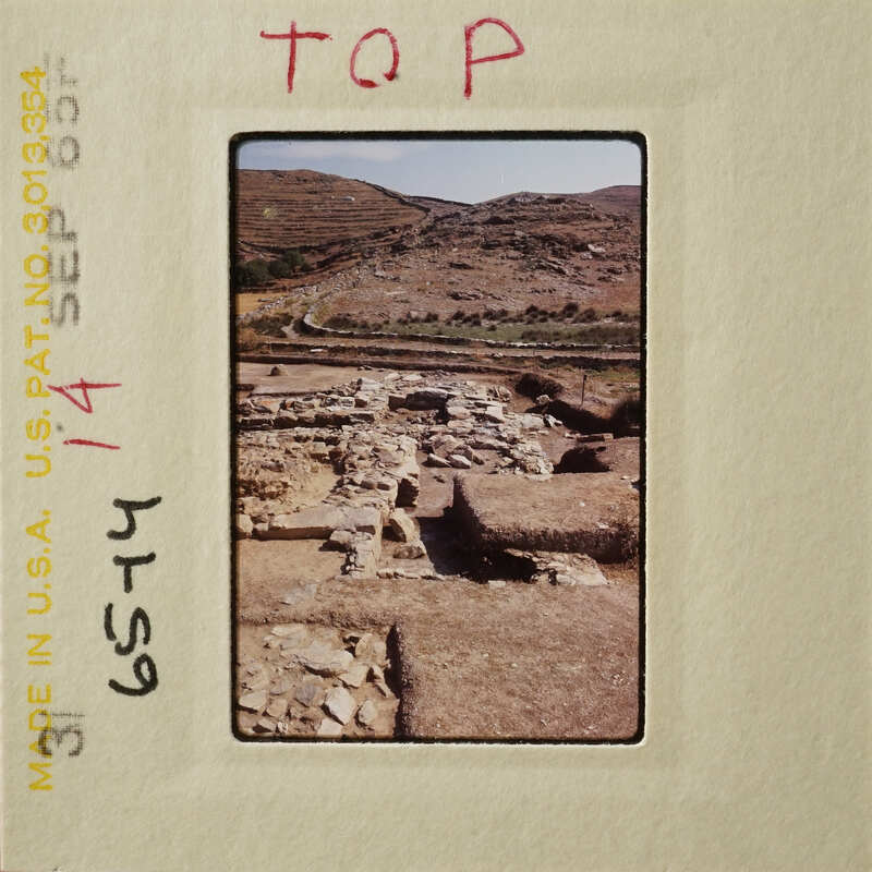 Ancient ruins with stone foundations and walls, set against a backdrop of rugged terrain and hills.