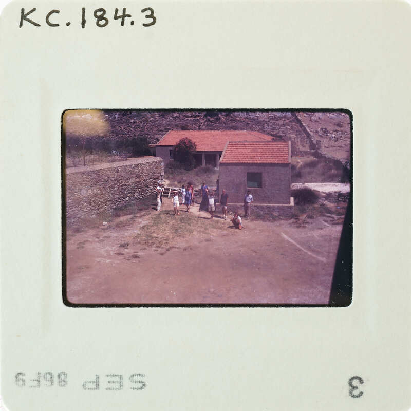 A vintage photograph of a group of people standing outside a small stone building with a red roof, taken from an aerial perspective.
