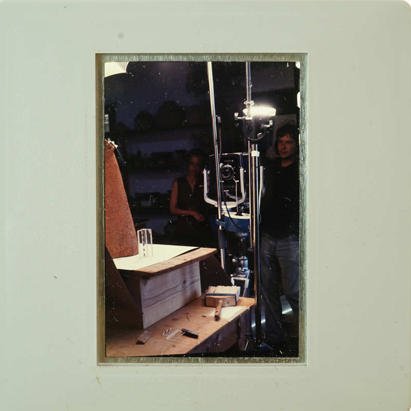 A man standing behind a camera on a tripod, with another person nearby and various items on a table.