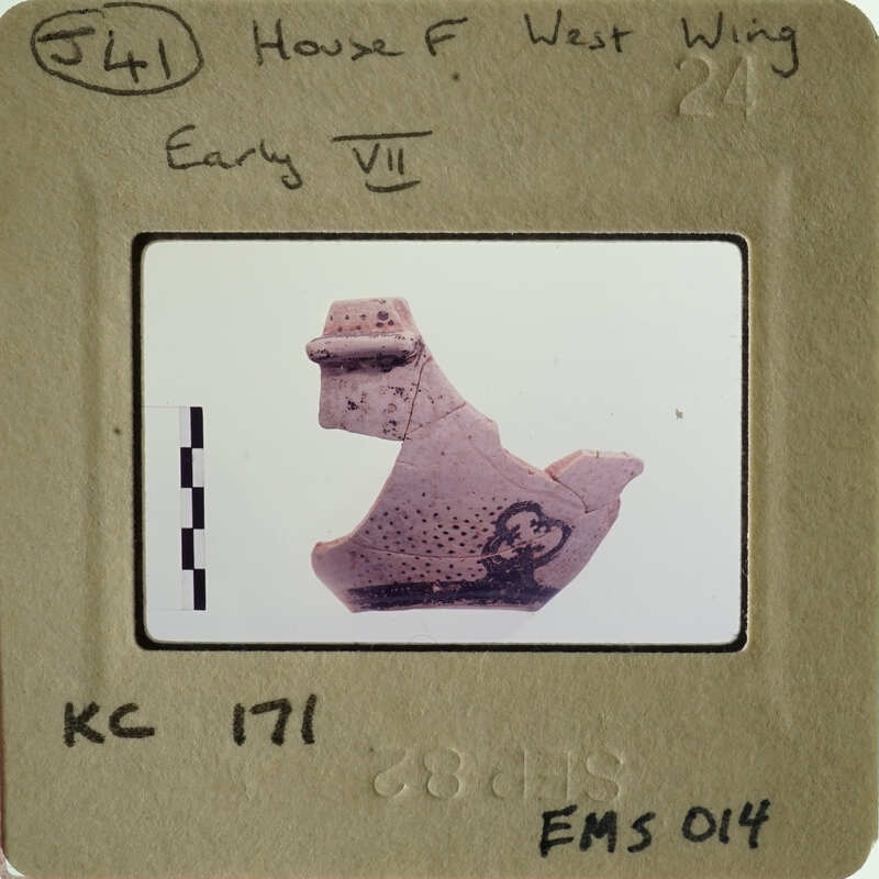 Ancient artifact on display, possibly a clay figurine with markings, photographed against a white background.