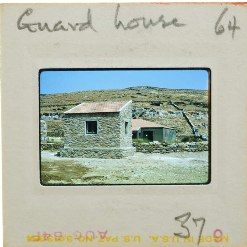 Old stone house with a red roof, surrounded by rock walls and mountains in the background.