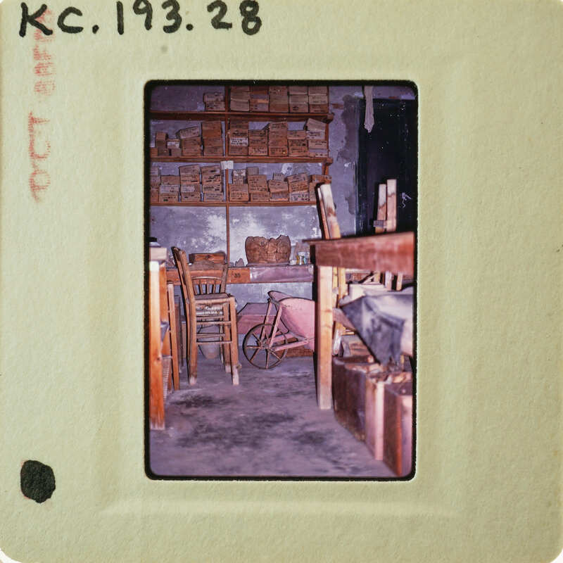 A vintage photograph of a rustic room with shelves, chairs, and a wheelbarrow.