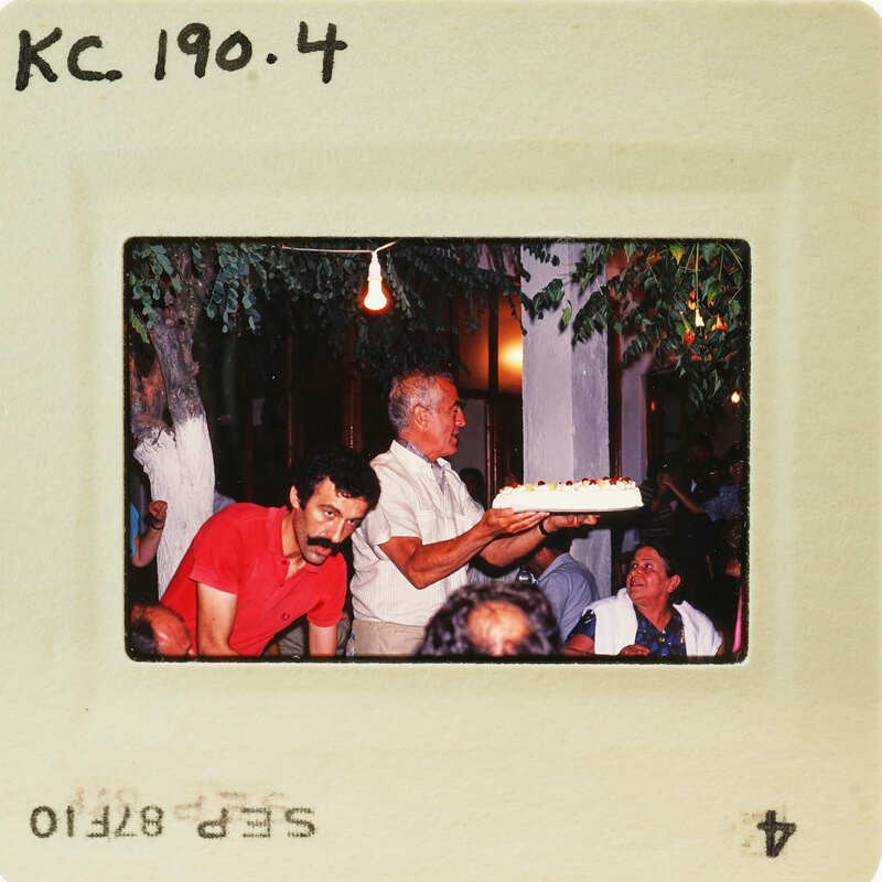 A vintage photo of a man holding a cake with people around him, possibly at a celebration or gathering. Catalog number KC.190.4