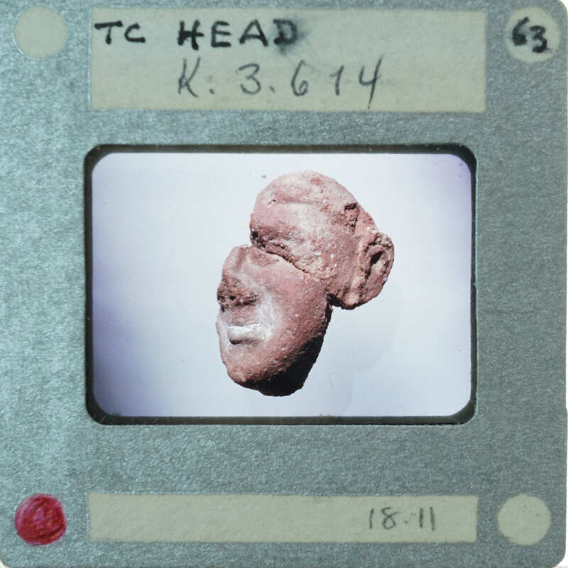 A close-up photograph of a small red figurine with a face, displayed on a white background.