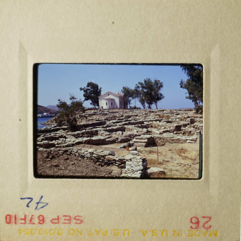The photo shows an ancient Greek temple on a rocky hillside, with a clear blue sky and a few trees in the background.