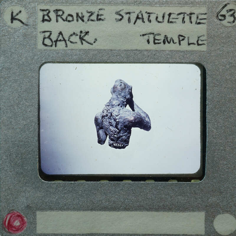 Small bronze statue of a figure with hands raised, from behind displayed on a white surface.