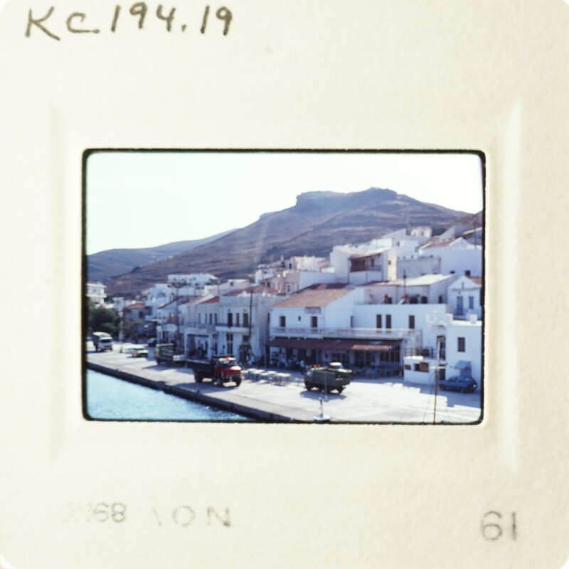 A black and white photograph of a coastal town with white buildings, taken from a distance.