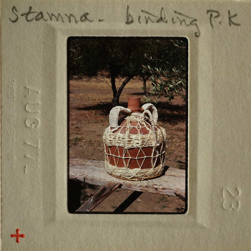An old-fashioned clay jug with woven handles and body, sitting on a wooden surface outdoors.