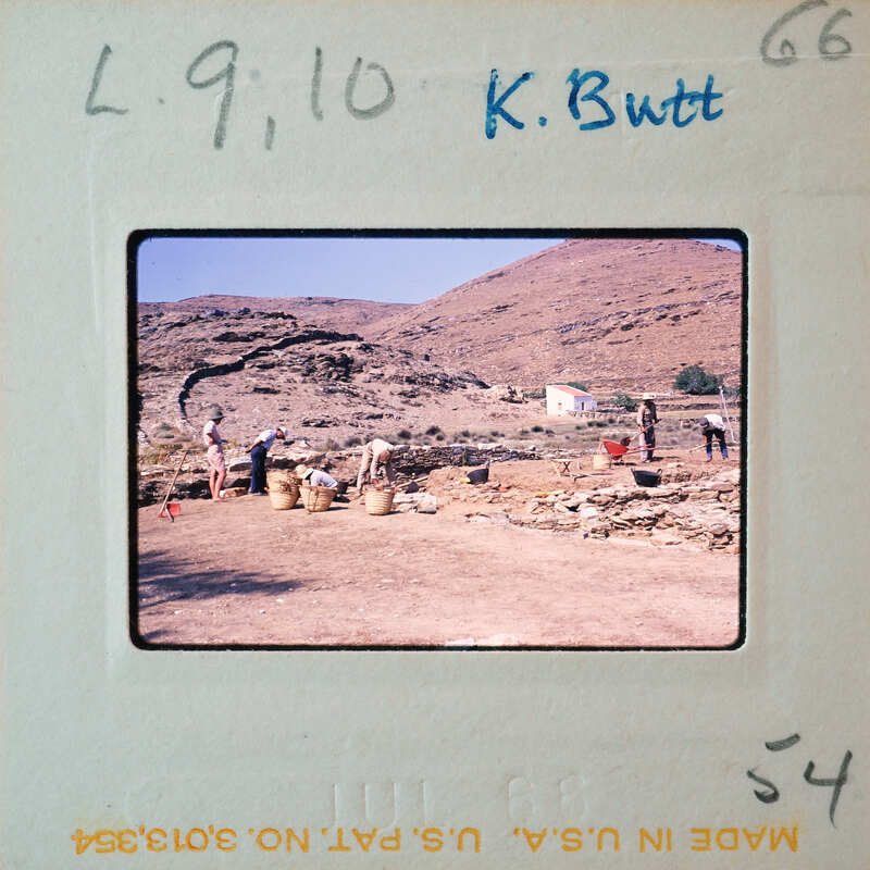 A vintage photograph of a group of people working on an archaeological site with stone ruins and mountains in the background.