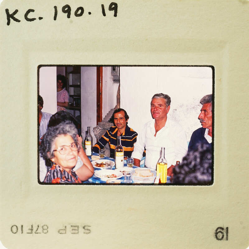 A vintage photo of a group of people sitting at a table, with an aged appearance and handwritten details on the bottom. Catalog number KC.190.19