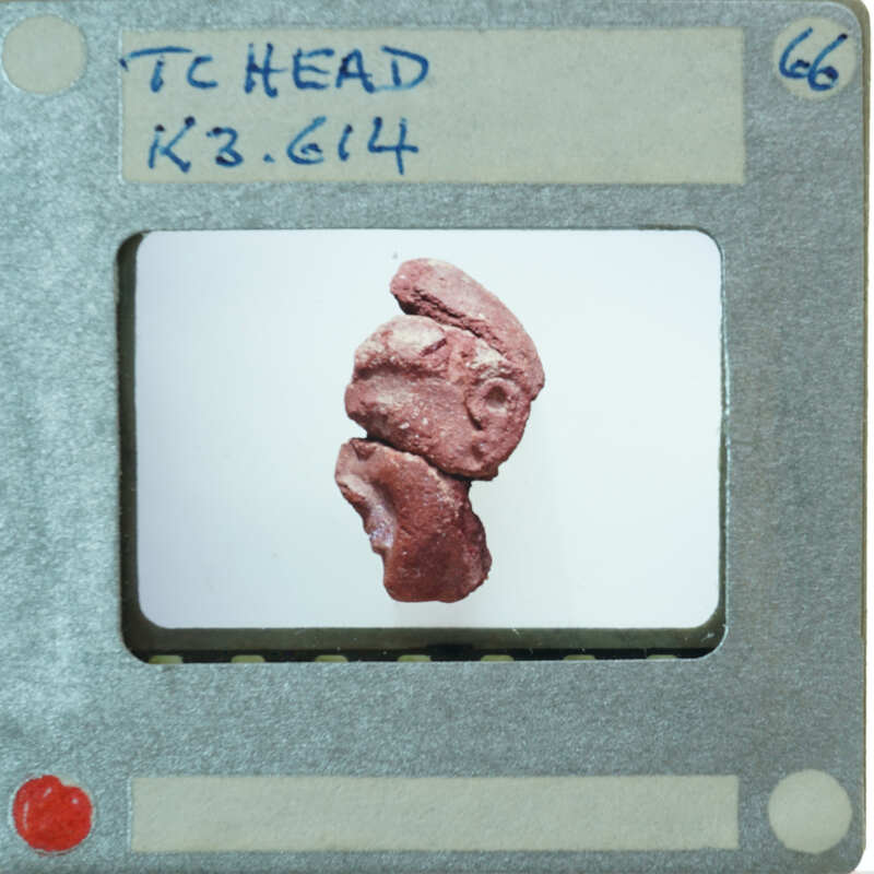 A close-up photograph of a small red figurine with a face, displayed on a white background.