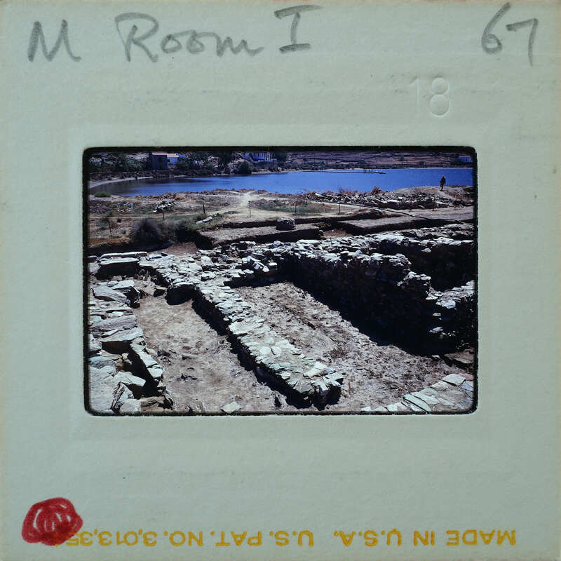 An old, partially excavated archaeological site with stone ruins and a body of water in the background.