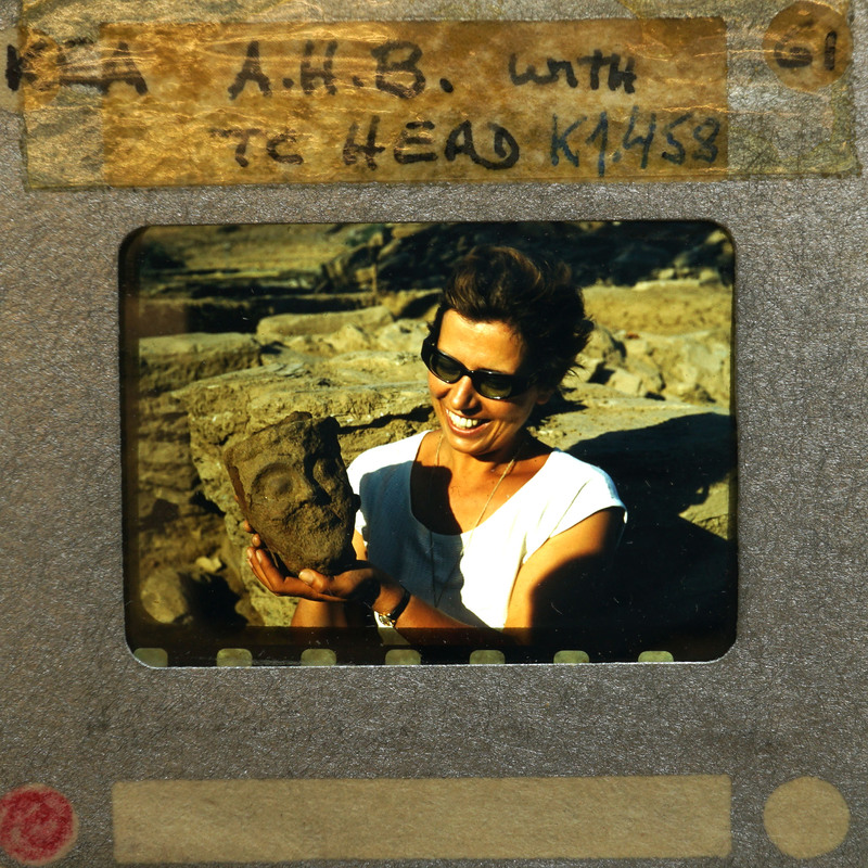 Woman holding terracotta artifact (K1.458) with smile, set against rocky background.