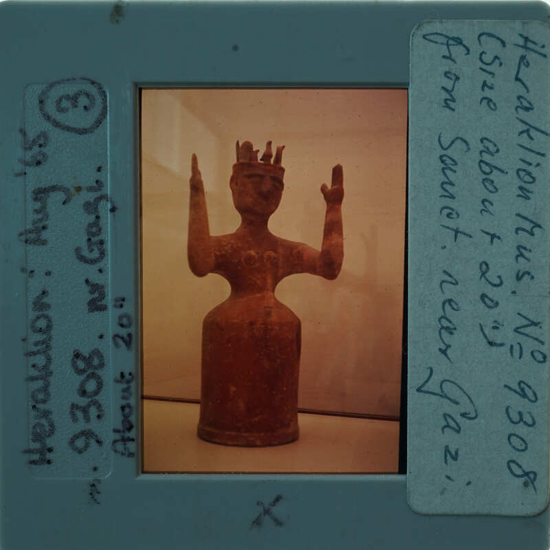 A clay figurine of a person with raised hands, displayed behind glass.