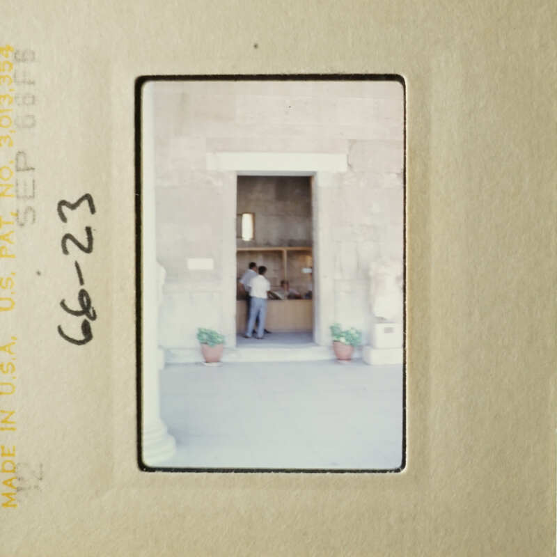 This is an old photograph of a person standing outside a building, possibly at a doorway or entrance, with another person inside.