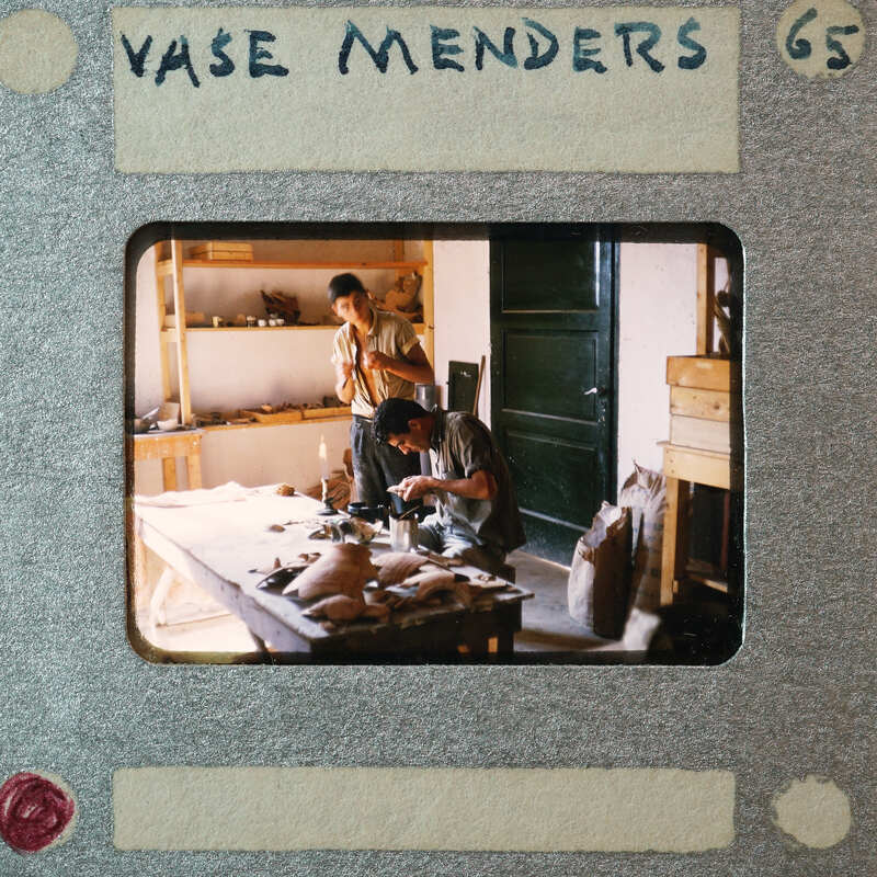 Two men working on pottery at a table with various tools and materials.