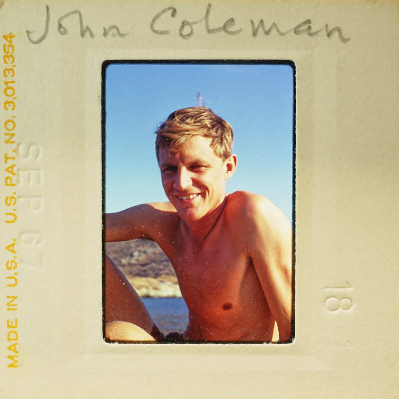A vintage Polaroid photo of a smiling young man with blonde hair, shirtless and sitting by the water.
