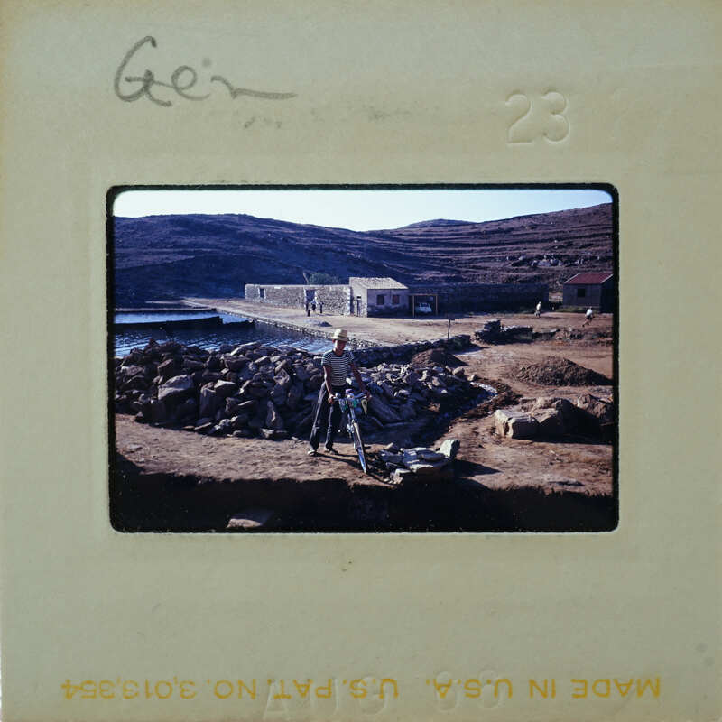 A person standing on a rocky shore near the sea, with a small village and mountains in the background.