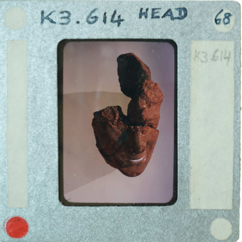 A close-up photograph of a small red figurine with a face, displayed on a white background.