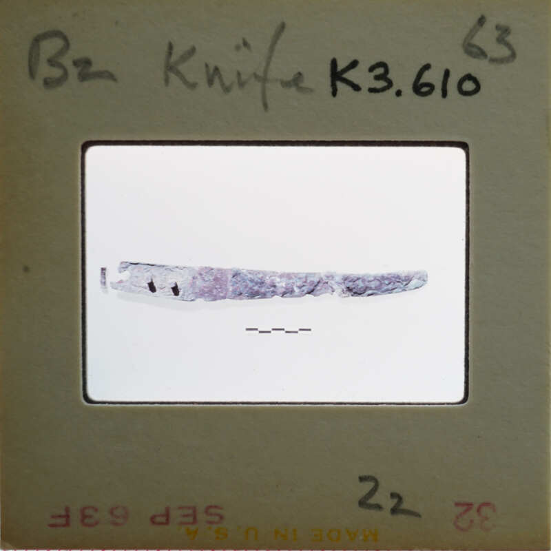 A small, curved metal object with a pointed end and a handle, possibly a knife or tool, on display in a case labeled "K360.610".