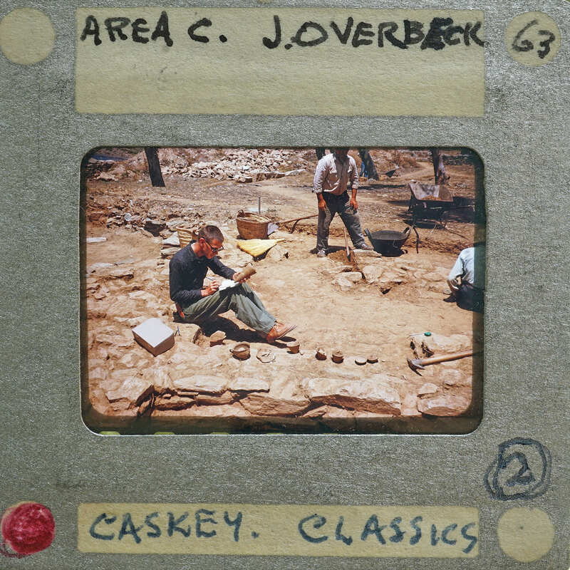 A vintage photo of a man sitting on the ground surrounded by rocks and other materials, with some people standing nearby, labeled as "Area C. Jovoberg 63" from Caskey Classics.