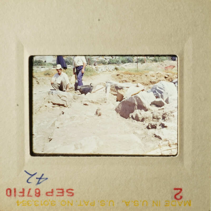 A vintage photograph showing two men working on an archaeological site with stone ruins and tools.